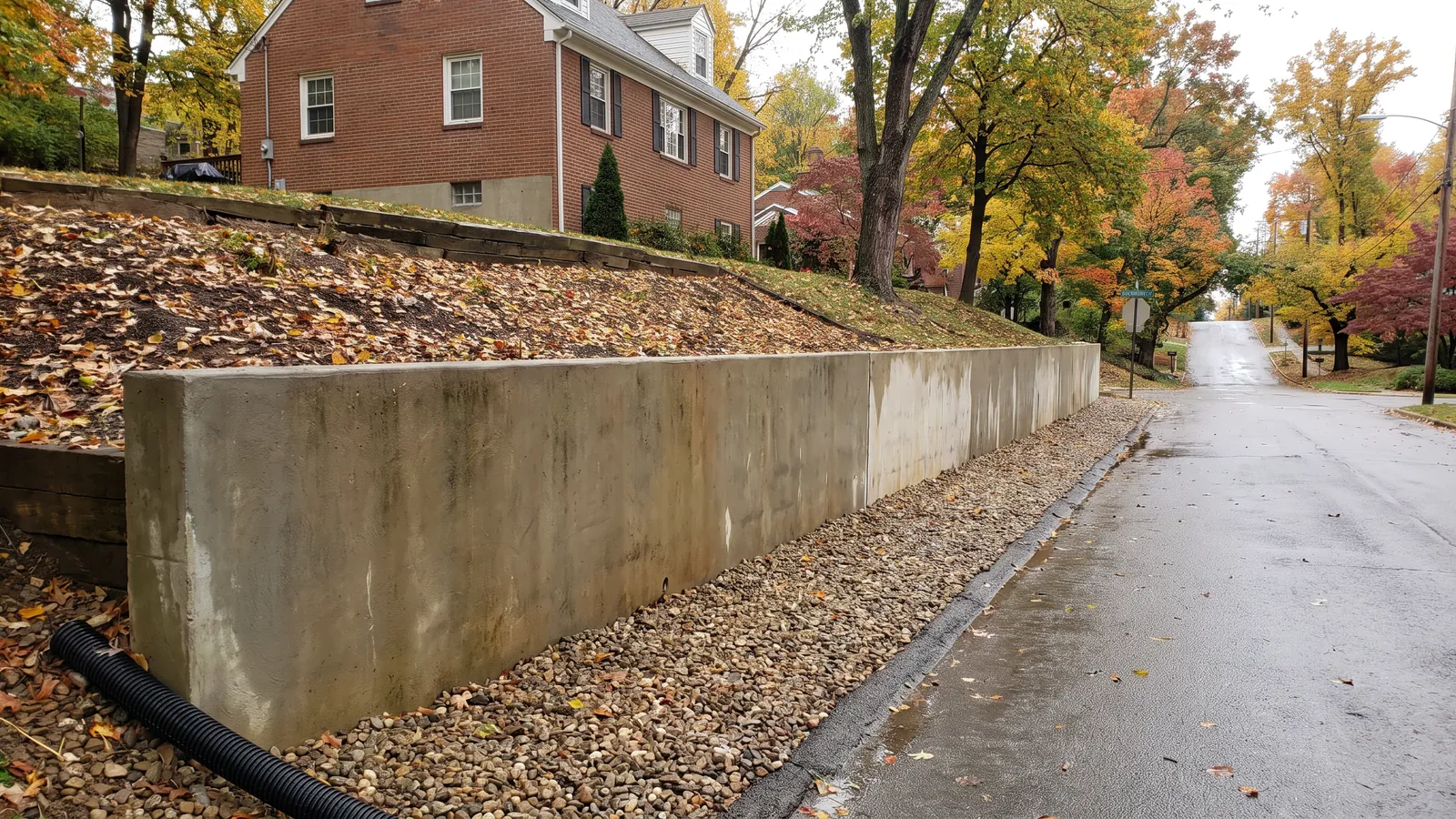 Poured concrete retaining wall holding a steep hillside lot below a brick house, drainage gravel and a drain pipe at the base