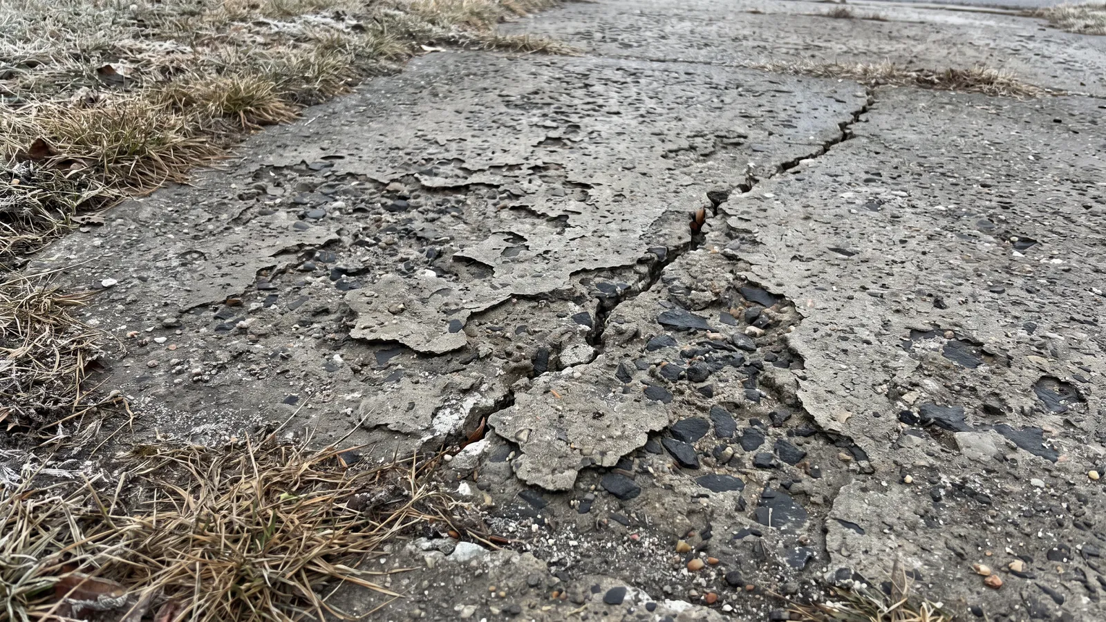 Residential concrete driveway surface spalled and pitted by winter road salt, flaking top layer exposing aggregate and a crack running through